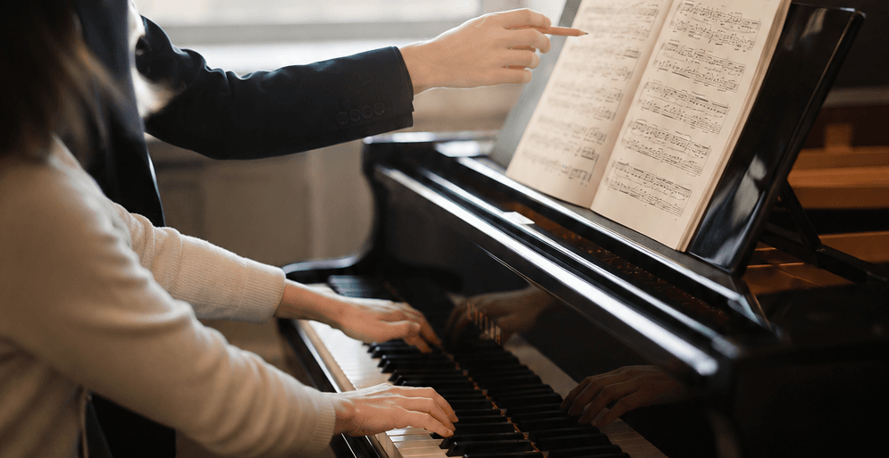 A piano teacher pointing to sheet music while a student plays a grand piano.